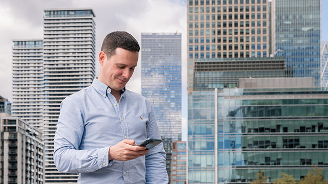 A smiling person looks at their phone with Canary Wharf skyscrapers in the background