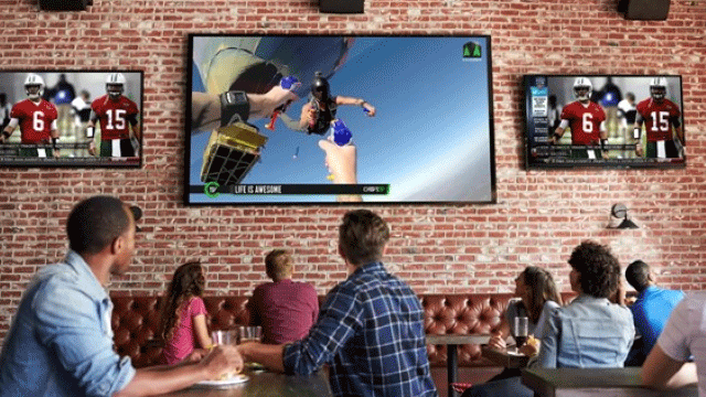 People in a bar watching sports and skydiving on multiple TVs mounted on a brick wall.