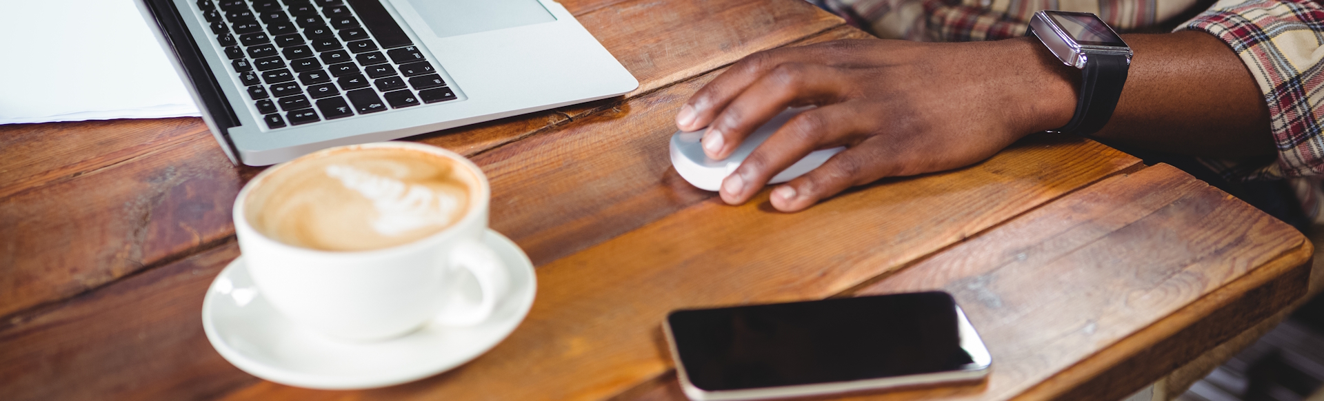 Wooden table with cup of coffee, mobile phone, laptop and someone's hand with a black watch holding the computer's mouse.