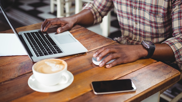 Wooden table with cup of coffee, mobile phone, laptop and someone with a black watch holding the computer's mouse.