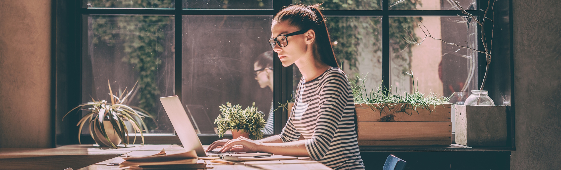 Woman in glasses, stripy top and ponytail working on a laptop on a wooden table by a big window.