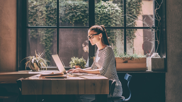 Woman in glasses, stripy top and ponytail working on a laptop on a wooden table by a big window.
