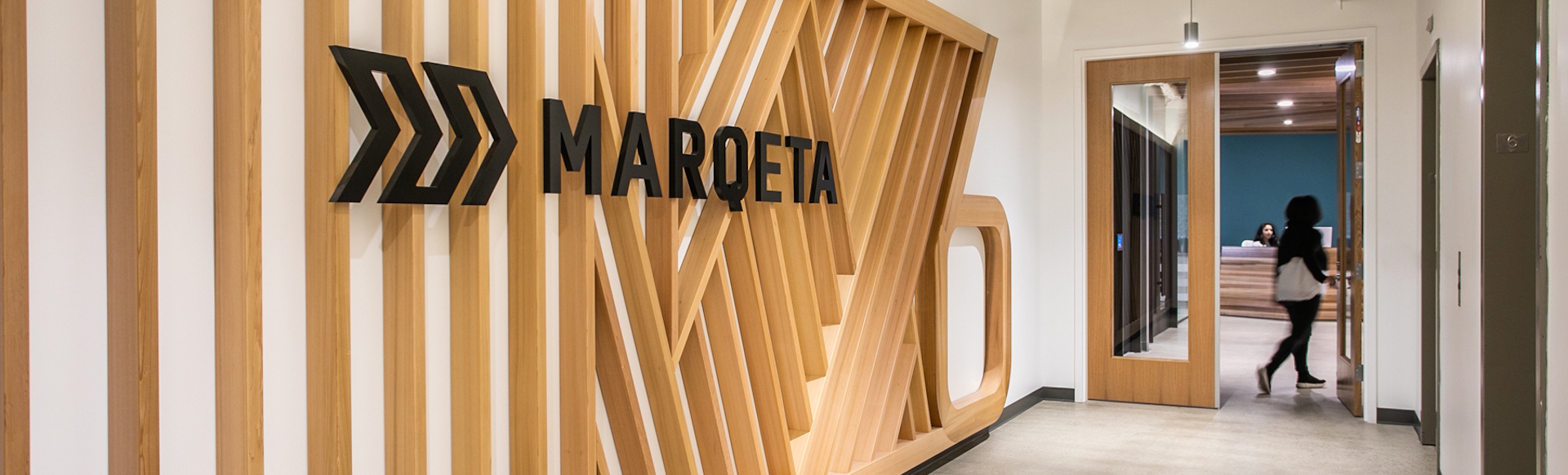 Marqeta office corridor with brand written in black capital letters on the wall and person walking into a room in the background.