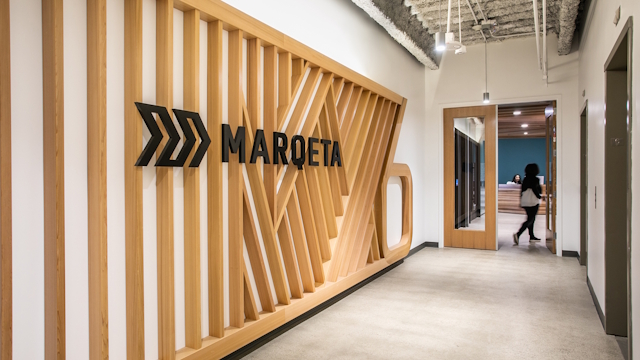 Marqeta office corridor with brand written in black capital letters on the wall and person walking into a room in the background.