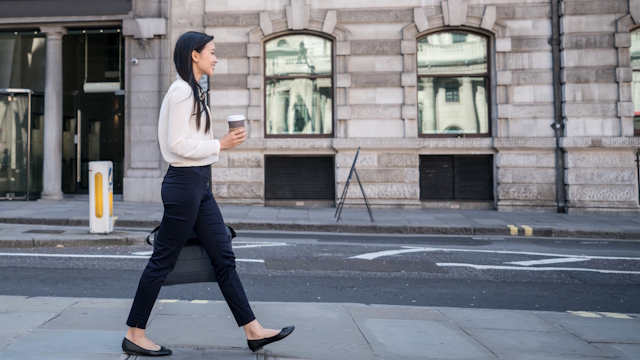 Asian woman dressed in black and white walking on London street with a cup of takeout coffee in her hand.