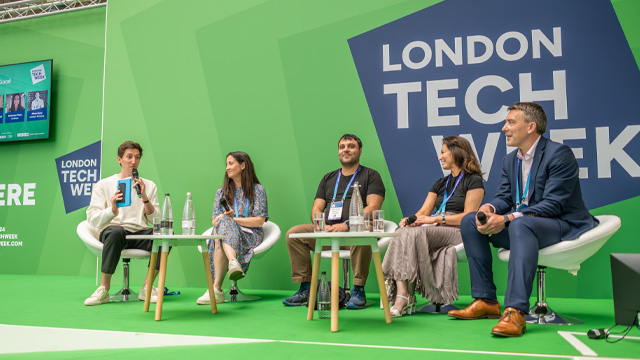 A view of a business conference with a room full of seated people looking at a stage with panelists.