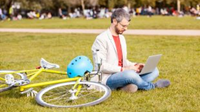 Man working on his laptop sitting on the grass at a park next to his bike