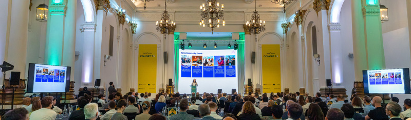 A large room filled with people attending a business conference looking towards a stage with a person speaking on a microphone.