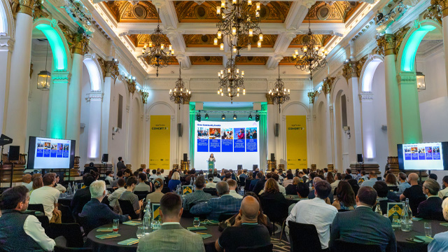 A large room filled with people attending a business conference looking towards a stage with a person speaking on a microphone.