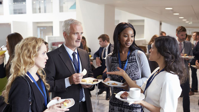 Business professionals networking at a corporate event in a modern venue, holding plates of food and engaging in conversation.