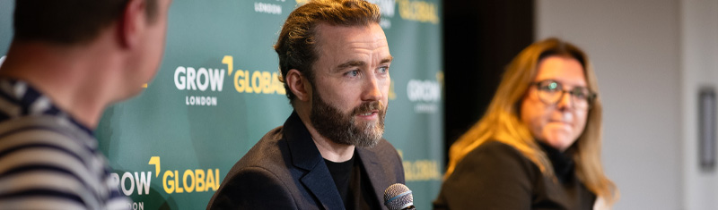A man speaking to an audience at a business event, with two other speakers at his sides listening.