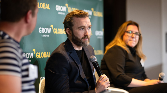 A man speaking to an audience at a business event, with two other speakers at his sides listening.