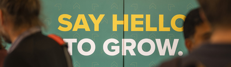 An image of people out of focus in the foreground with words on a wall that read "say hello to Grow"