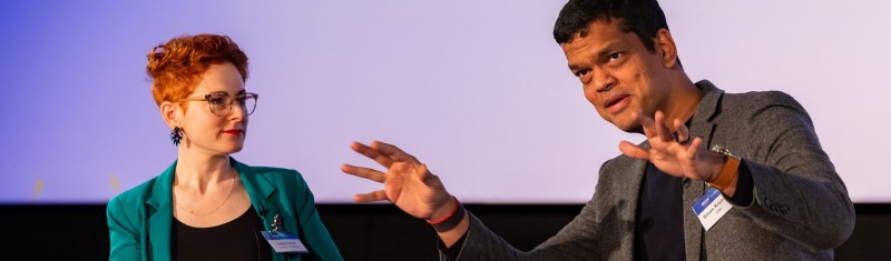 A man speaking at a business conference making animated gestured with his hands while a woman next to him listens attentively.