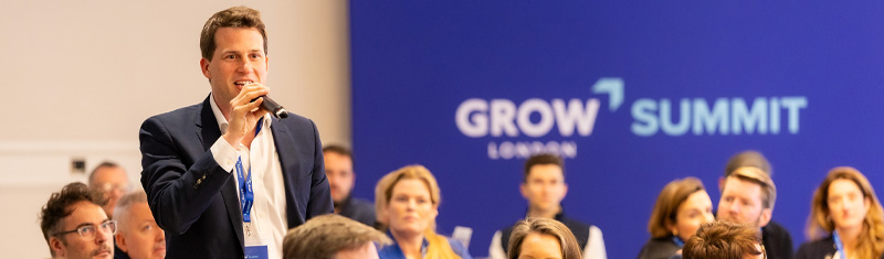 A man wearing a suit holding a microphone speaking in a business event in a room full of people.