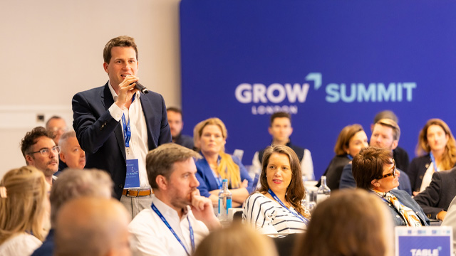 A man wearing a suit holding a microphone speaking in a business event in a room full of people.