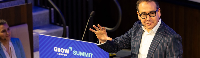 A man wearing a dark suite is standing at a lectern speaking to an audience at a business event. The lectern is branded with a blue colour and reads Grow Summit.