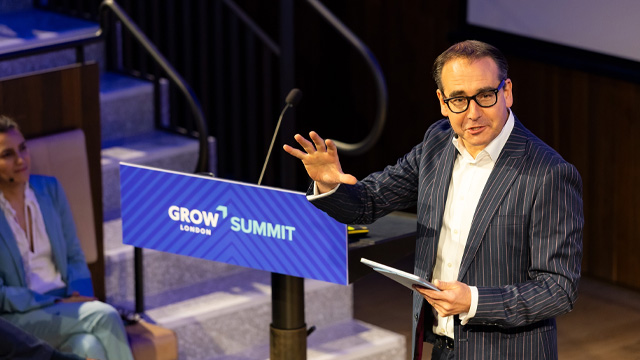 A man wearing a dark suit is standing at a lectern speaking to an audience at a business event. The lectern is branded with a blue colour and reads Grow Summit.