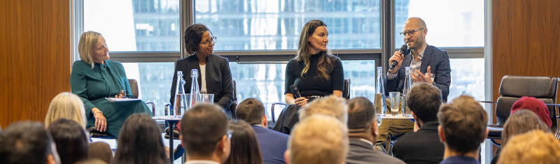 A panelist speaking to a crowd of people in a business conference with cityscape views in the background.