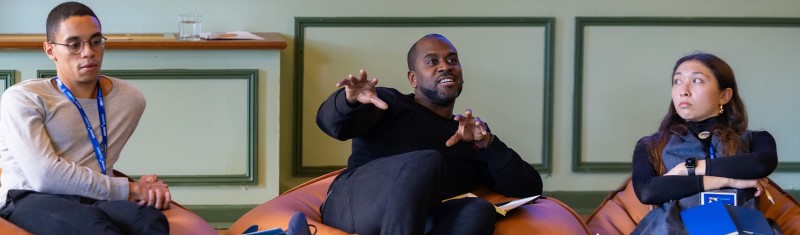A man speaking at a business conference making gestures with his hands, while two other panelists listen.