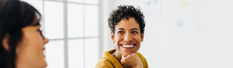 Two women sitting at a desk in a brightly lit, modern office, smiling and talking. 
