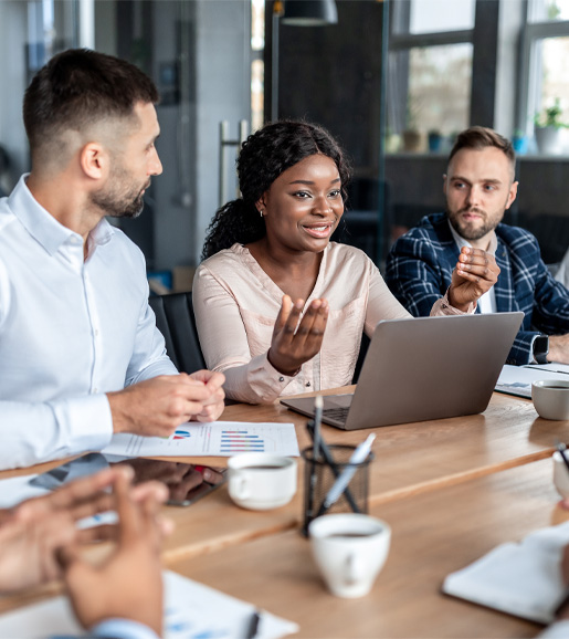 People sitting at a meeting table in a modern office, a woman is speaking and smiling and the people are listening. 