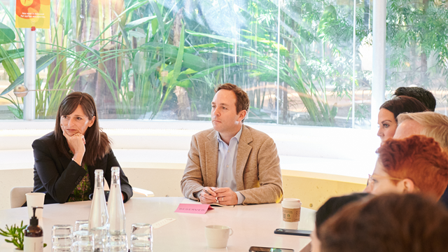 People sitting at a round table in a modern conference room, attentively listening during a meeting, large green plants visible outside.