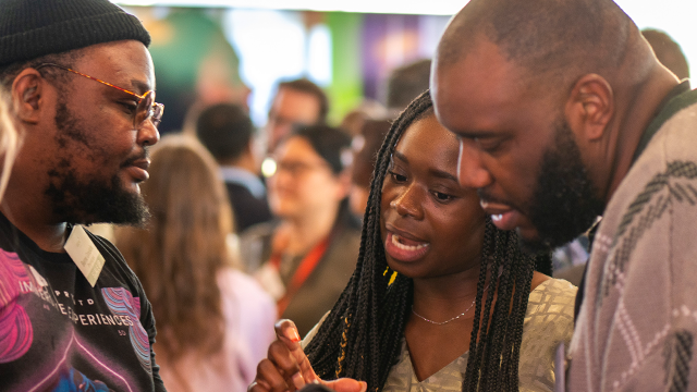 A group of three people engage in a discussion at a networking event, with a blurred crowd in the background.