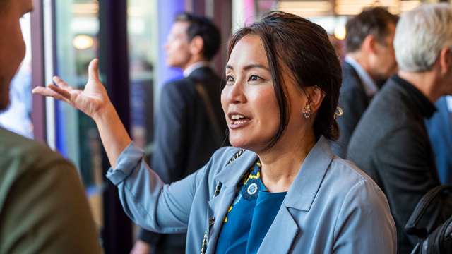 A woman in business attire engages in conversation at a networking event, with several people in the background.