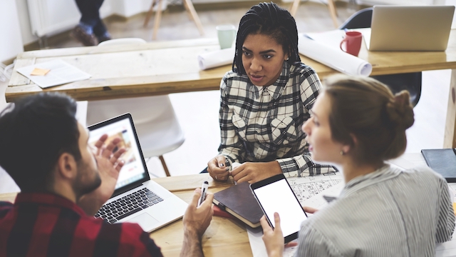 Three people having a meeting in an office at a table with a computer, phone and notebooks.