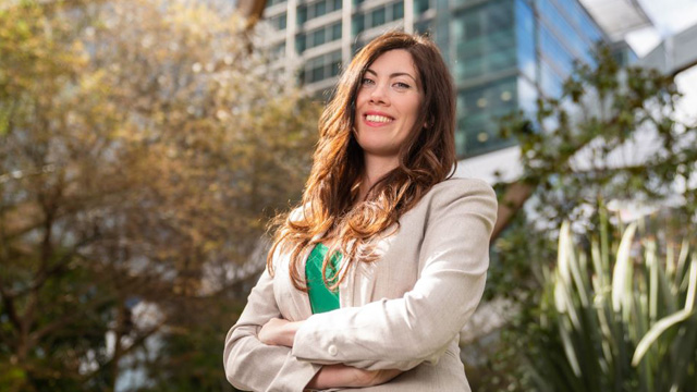 Smiling professional woman in a blazer stands confidently in a city park with modern buildings in the background.