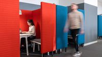A woman working in an office booth and a man walking past.