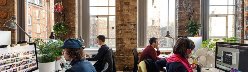 Four people working on computers with large windows in the background.