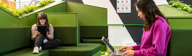 Two women sitting on green sofas reading and working on a laptop and tablet.