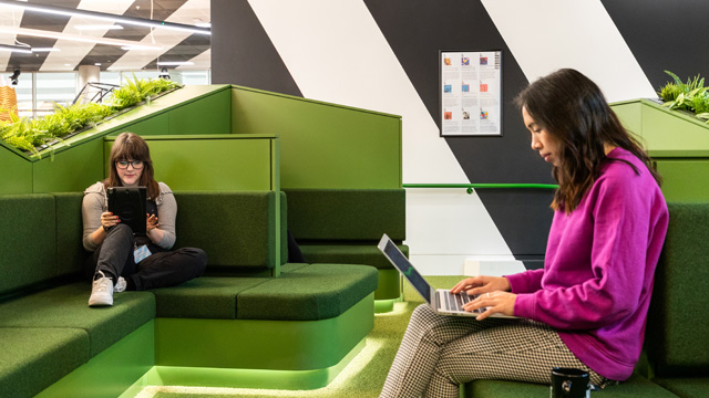 Two women sitting on green sofas reading and working on a laptop and tablet.