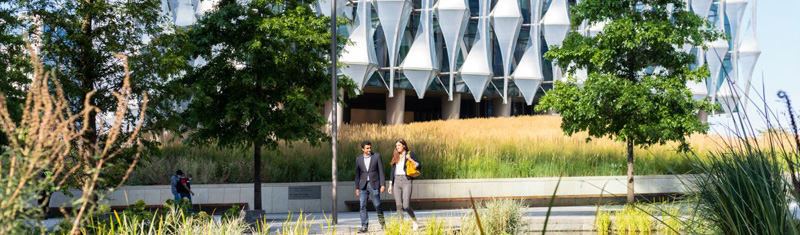 Two people walking in a garden with a modern building in the background.