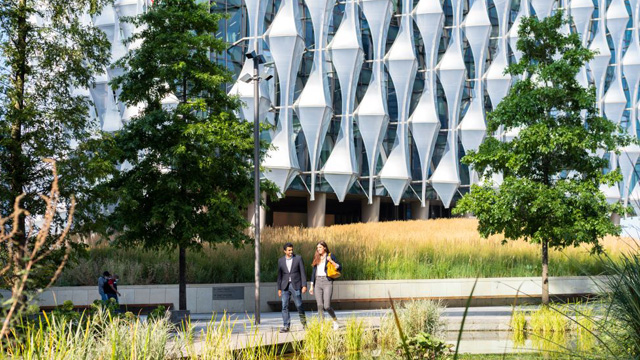 Two people walking in a garden with a modern building in the background.