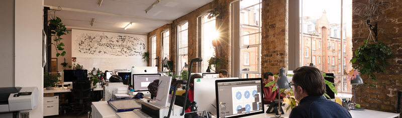 A contemporary office space with exposed brick walls and floor-to-ceiling windows, with people working at desks.