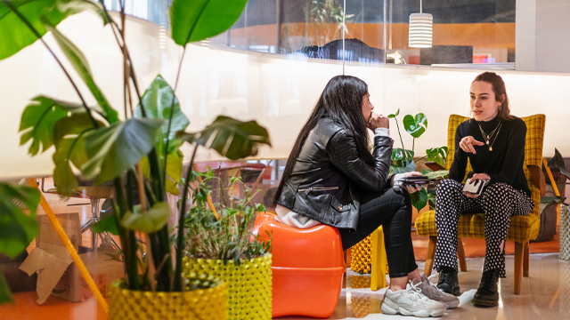 Two women talking and sitting on chairs in a colourful office space.