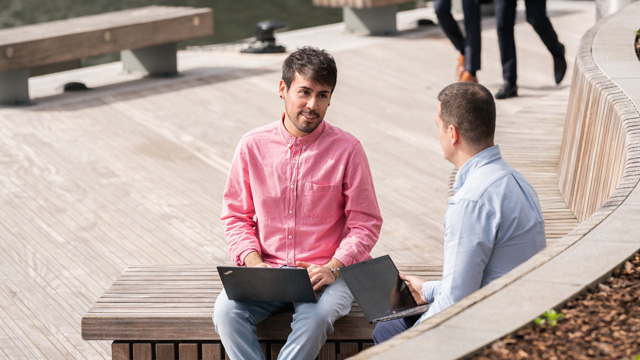 Two men talking and sitting outdoors in Canary Wharf next to the river Thames.